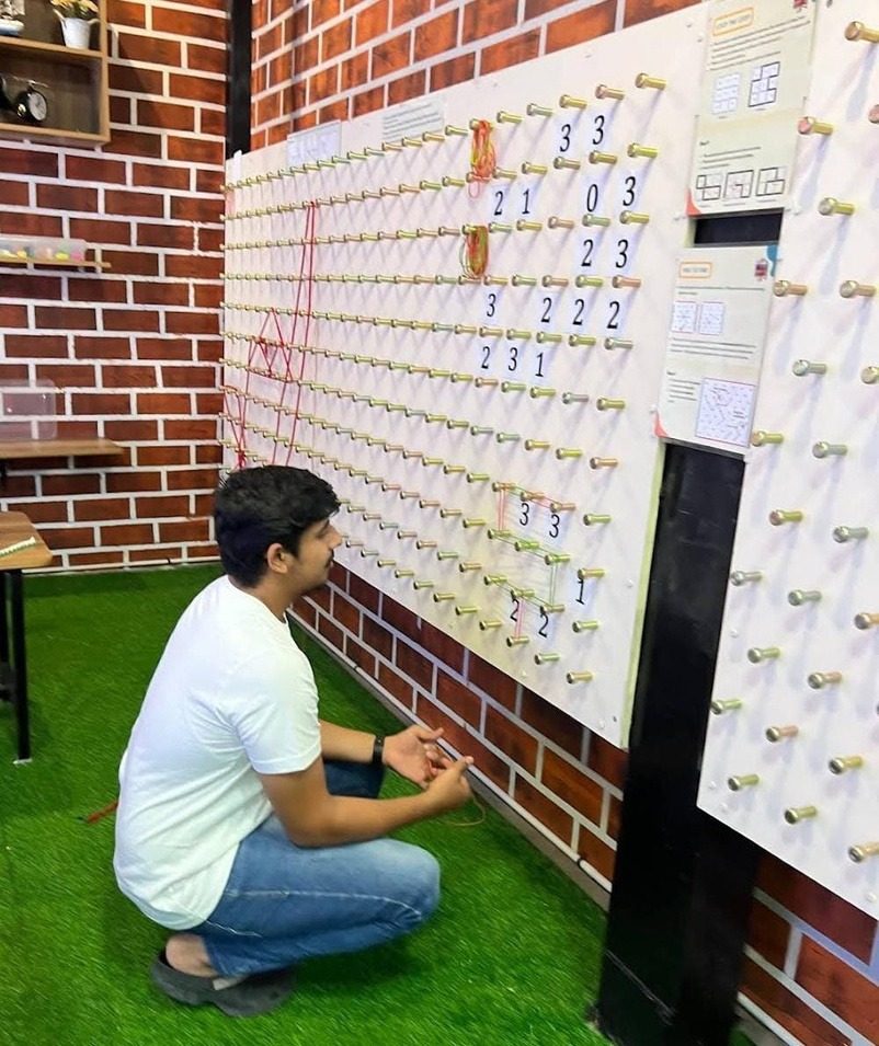 A visitor kneeling at the geoboard wall, stretching bands between pegs to make shapes